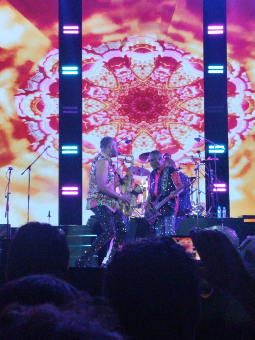 Kool and the Gang sur scène, lors d'un concert. saxophone et une basse, devant un grand écran affichant des motifs lumineux et colorés, photo gratuite