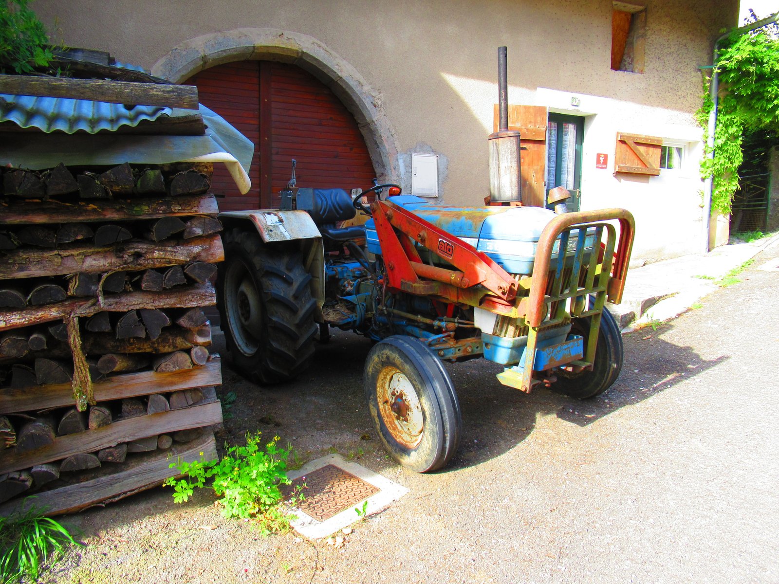 Un tracteur agricole garé près d'un bâtiment, à côté d'une pile de bois de chauffage