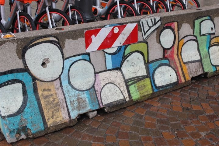 Barrière en béton décorée de graffitis, représentant des personnages stylisés ou des visages. On y voit également un panneau de signalisation rouge et blanc à rayures, et des vélos en arrière-plan, photo gratuite