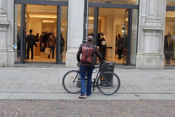 Une personne debout, à côté d'un vélo noir, attaché à un poteau. La personne porte un sac à dos marron, il est devant une galerie commerciale, photo gratuite