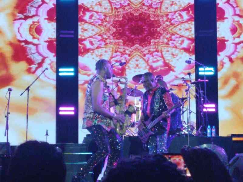 Kool and the Gang sur scène, lors d'un concert. saxophone et une basse, devant un grand écran affichant des motifs lumineux et colorés, photo gratuite